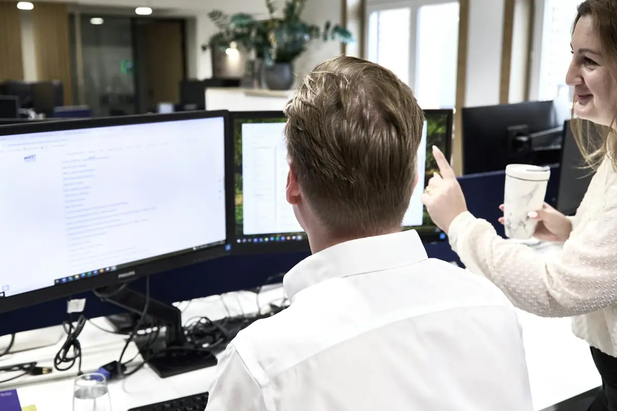 Twee mensen overleggen bij een beeldscherm op een kantoor met meerdere werkplekken